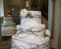 Three tiered cake covered in white fondant with beaded buttercream detail atop a tree trunk stand. Three white roses, one on each layer of cake, and decorative twigs surround the layers of cake. 