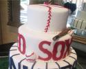 Three layers of cake stacked covered in white fondant with buttercream beaded detail. The design on the first layer, resembling a baseball, is made out of red buttercream. The letters and decorative pieces on the second and third layers are all made out of red and blue fondant.