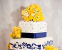 Three square tiers of cake stacked and covered in white fondant. Each layer is surrounded by a metallic blue ribbon. Layers one and three of cake are dotted with blue buttercream, the second layer has a beautiful flower design painted on all four sides. Get creative with your cake setup! Yellow and white flowers add color and brightness to the look.
