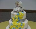 Three round tears of cake stacked and covered in white fondant. The layers have a gray ribbon going around with small blue and yellow flowers made out of fondant. The cake topper is a cartoon rendition of the bride and groom. 