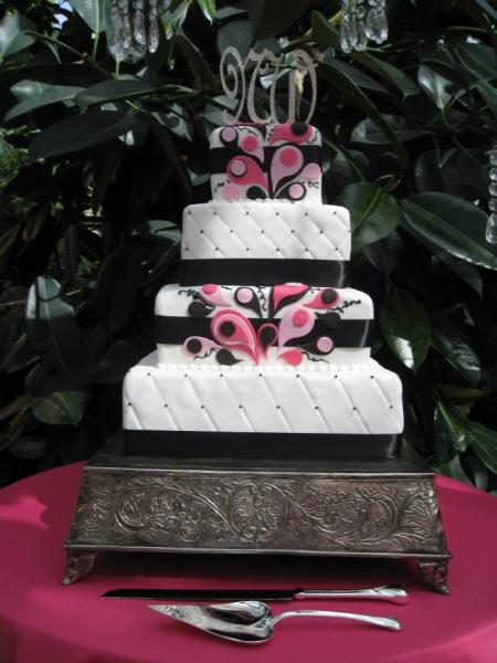 Square layers of cake stacked atop a metal-looking stand to create the classic wedding cake look. All layers are covered in white fondant with a black ribbon going around each cake. The first and third layers have a design made out of fondant in different shades of pink. This adds a pop of color to a simple yet elegant cake!