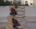 Beautiful three tiered cake covered in white fondant on top of a white stand. The three layers of cake are decorated with swirling stripes of taupe colored fondant and flowers that are also made out of blue, green and red fondant.