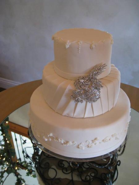 Three tiered cake covered in white fondant with buttercream details on the first and third layers. The second layer sits a silver decorative piece, adding the cake a touch of class and elegance.