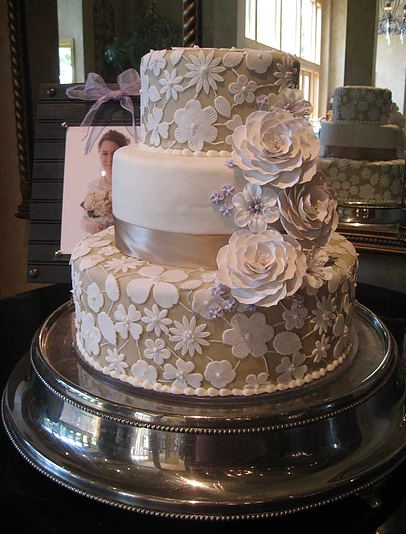 Three tiered cake covered in white and taupe fondant. You can see a flower pattern that resembles lace on the first and third layers. The flowers on the right side are also made out of fondant. 